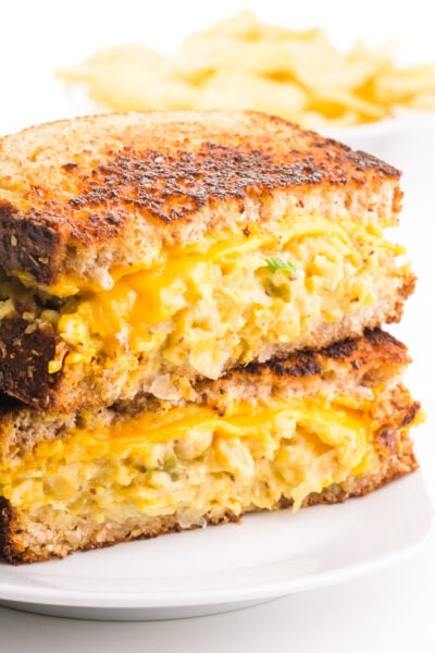 Two halves of a sandwich are stacked, revealing vegan tuna salad and melted vegan cheese on golden grilled bread. There are potato chips in the background.