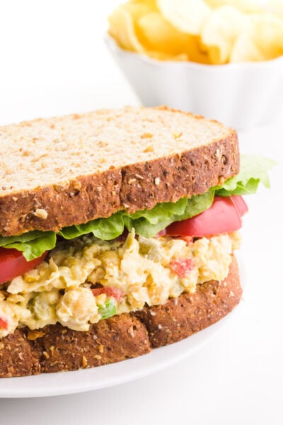 A vegan tuna salad sandwich has lettuce and sliced tomatoes on it. There's a bowl of potato chips in the background.
