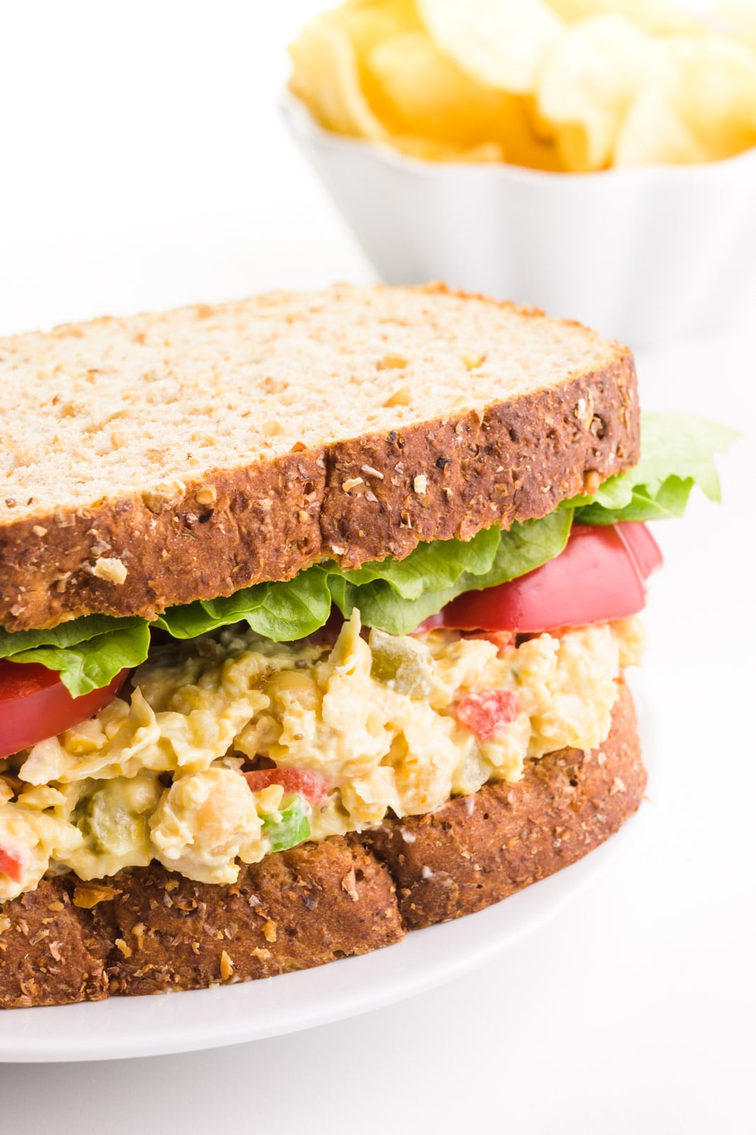 A vegan tuna salad sandwich has lettuce and sliced tomatoes on it. There's a bowl of potato chips in the background.