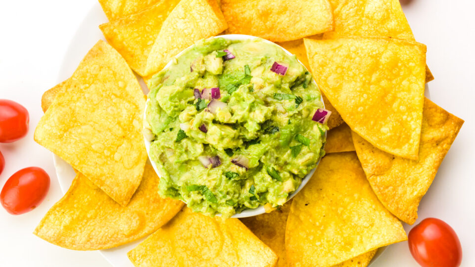 Looking down on a bowl of guacamole surrounded by air fried tortilla chips. There are cherry tomatoes and fresh limes nearby.