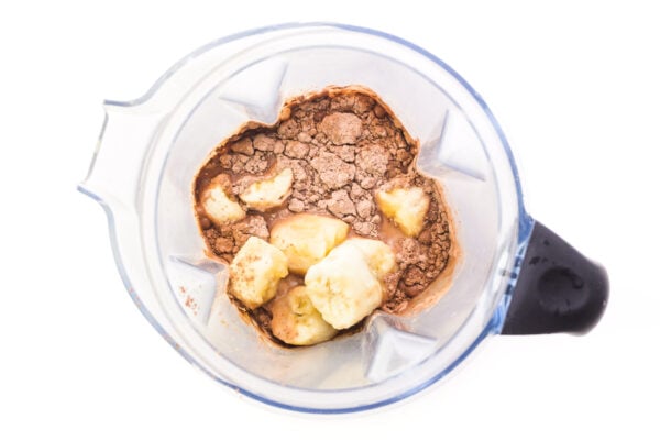 Looking down on a blender jar with ingredients for a smoothie, such as chocolate protein powder and banana chunks.