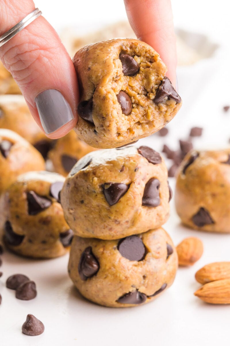 A hand holds an almond butter ball, hovering over a stack of two. There are more balls, raw almonds, and chocolate chips around it.