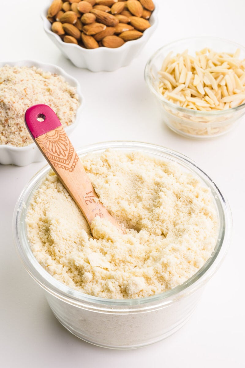 A wooden measuring spoon is in a bowl of almond flour. There are bowls in the background holding almond meal, raw almonds, and almond slivers.