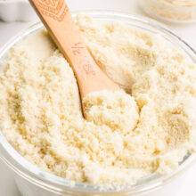 A closeup of a wooden measuring spoon in a bowl of almond flour. There are two bowls in the background holding almond meal and almond slivers.