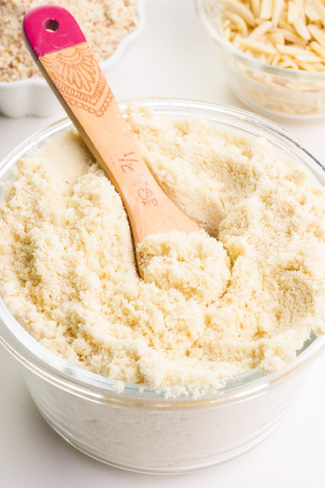 A closeup of a wooden measuring spoon in a bowl of almond flour. There are two bowls in the background holding almond meal and almond slivers.