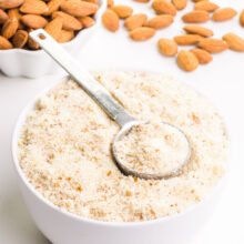 A metal measuring spoon sits in a bowl full of almond meal flour. There's a bowl of almonds in the background with more almonds on the table beside it.