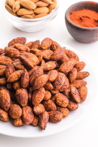 A plate holds a pile of bacon-flavored almonds. There is a bowl of raw almonds and a bowl with spices in the background.