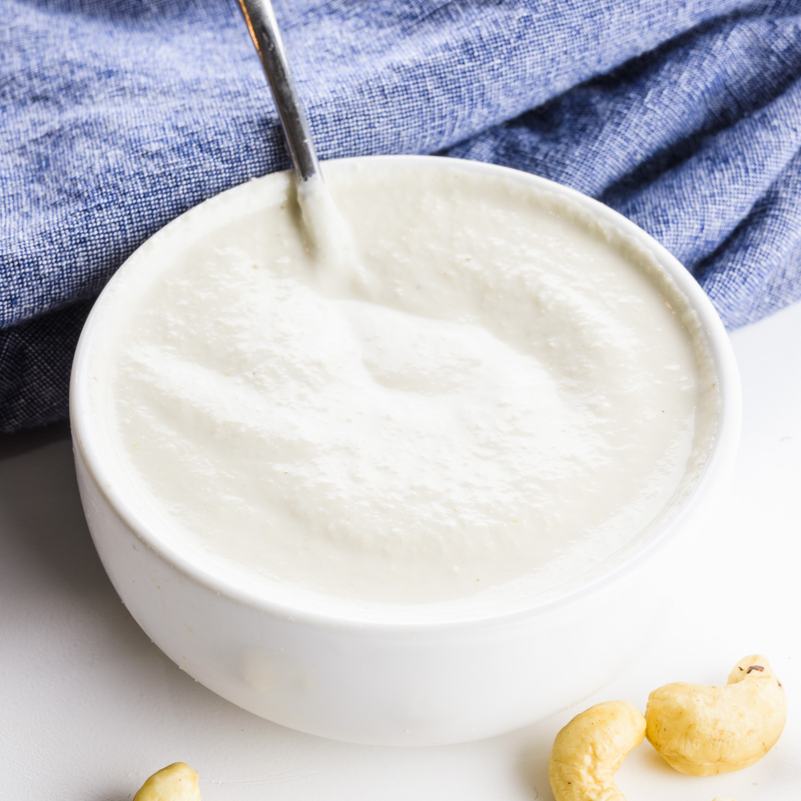 A white bowl holds cashew cream. There's a blue towel behind it and some cashews in front of it.