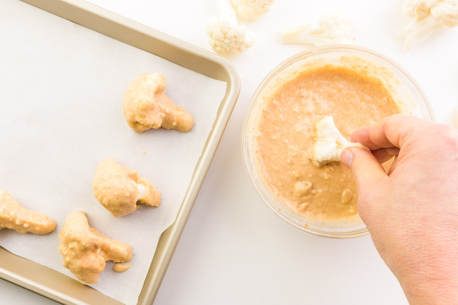 A hand holds a cauliflower floret dipping it in batter. There are other florets waiting to be dipped by the bowl. Some florets have already been dipped and are on a baking sheet.