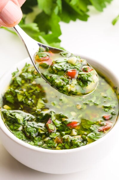 A hand holds a spoonful of chimichurri sauce, hovering over a bowl full of it. There are fresh green herbs in the background.