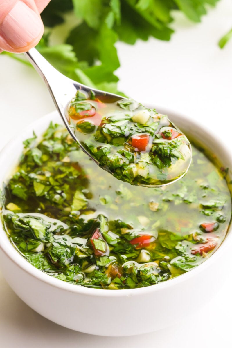 A hand holds a spoonful of chimichurri sauce, hovering over a bowl full of it. There are fresh green herbs in the background.