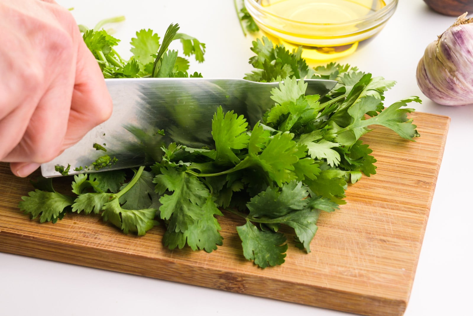 A hand holds a knife cutting cilantro on a wooden cutting board. There other ingredients in the background, such as a bowl of olive oil and garlic.