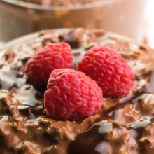 A bowl of chocolate overnight oats has raspberries on top. There's another bowl of oats the background.