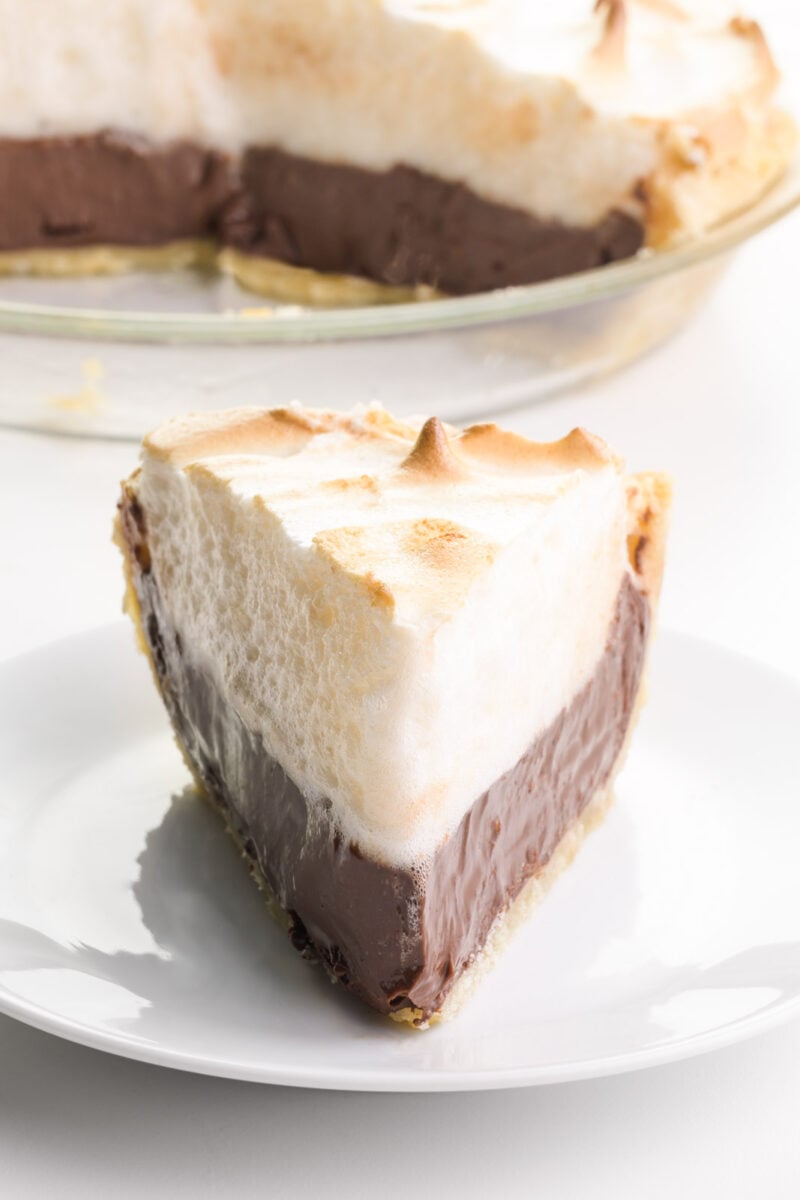 A slice of chocolate pie sits on a plate. It's piled high with meringue. The rest of the pie is in a glass pie pan behind it.
