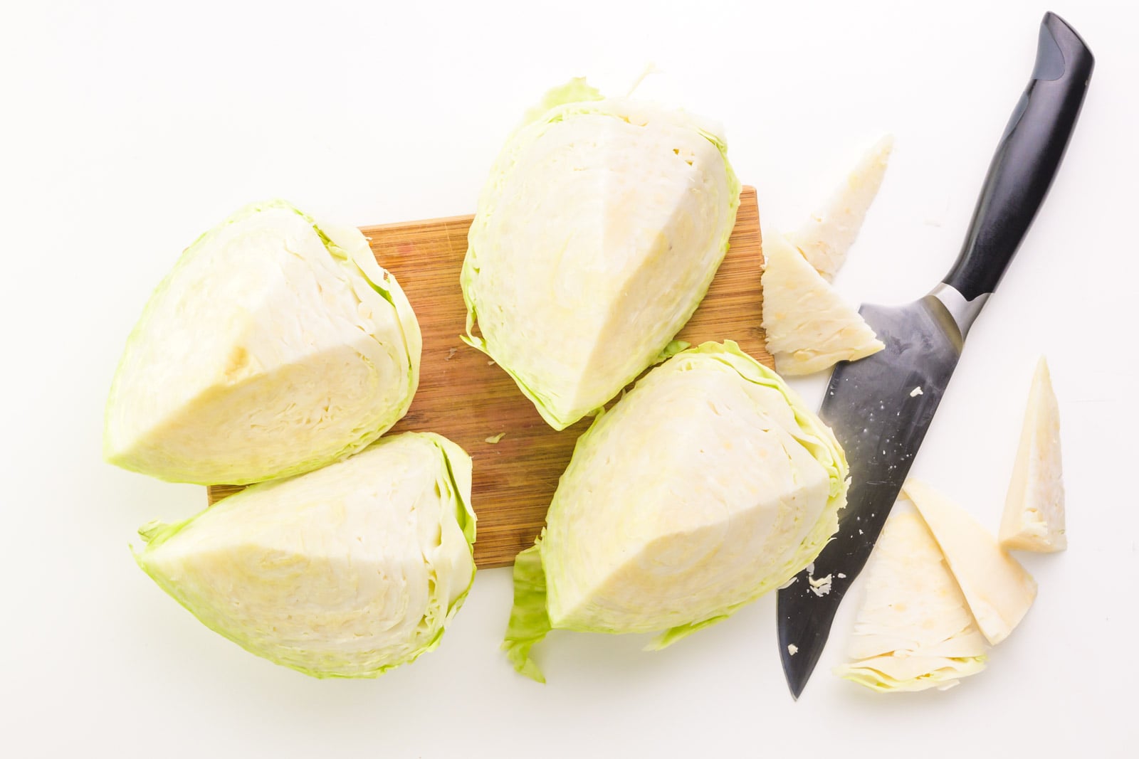 Looking down on a cutting board holding four quarters of green cabbage, each with the core cut out. There's a chef's knife beside it with the cut-out core beside it.