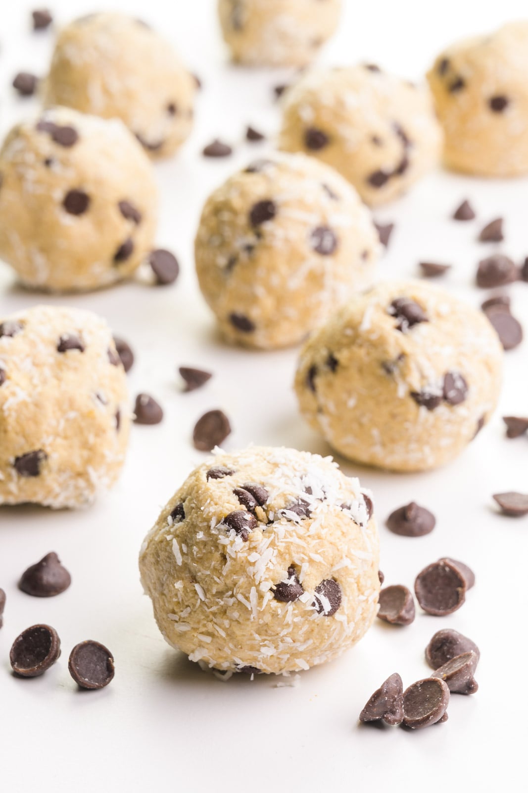 Several coconut balls are on a table surrounded by chocolate chips.