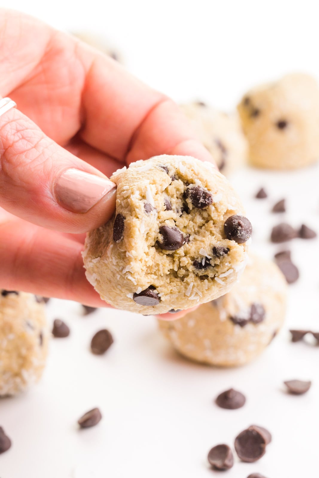 A hand holds a protein ball with a bite taken out of it, showcasing chocolate chips throughout. There are more balls and chocolate chips in the background.