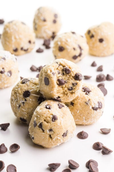 Coconut balls sit in a stack with a bite taken out of the top one. There are more balls and chocolate chips on the table and in the background.