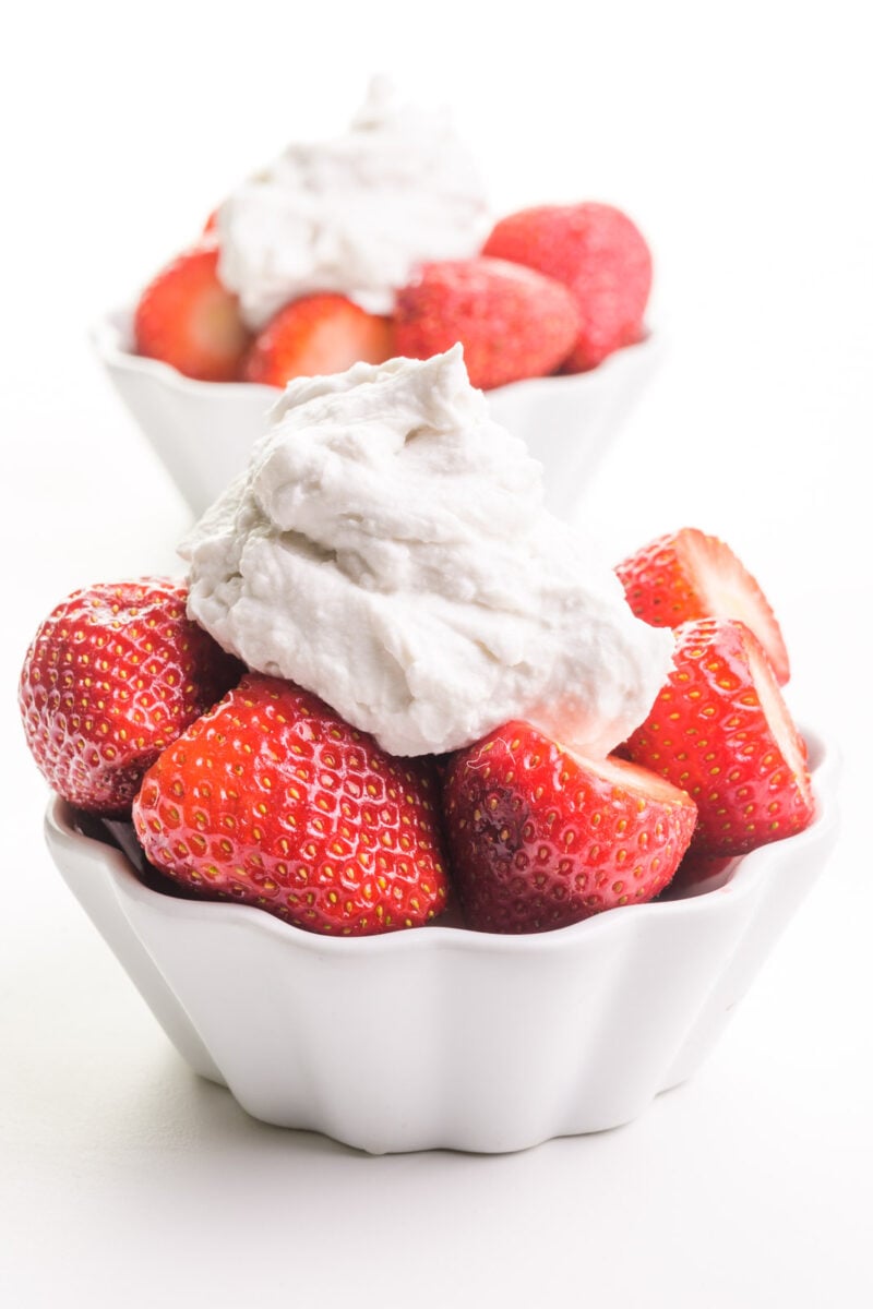 Two bowls of strawberries have coconut milk whipped cream on top. One is sitting in from the other.