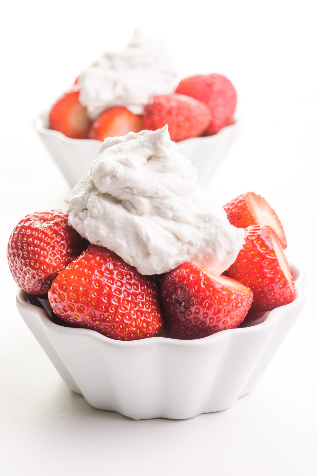 Two bowls of strawberries have coconut milk whipped cream on top. One is sitting in from the other.