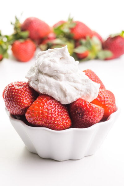 A bowl of strawberries has coconut whipped cream on top. There are more strawberries in the background.