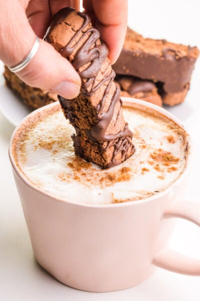 A hand holds a chocolate biscotti cookie and is dipping it in a pink mug with chai latte in it.