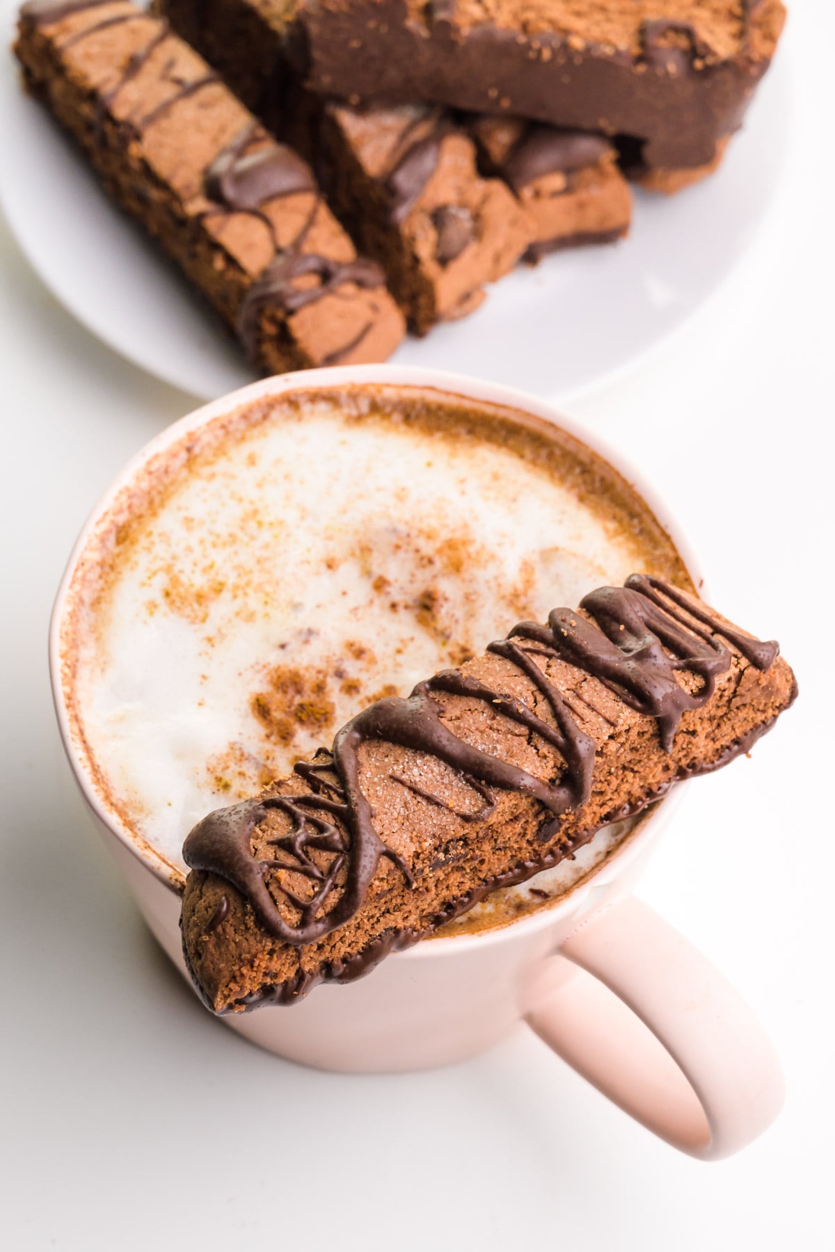 A biscotti sits on the edge of a mug full of hot tea. There's a place with more biscotti in the background.