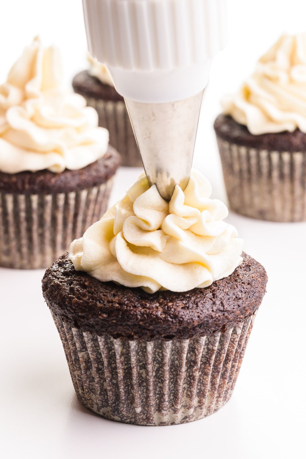 A piping bag is full of ermine frosting that is being piped onto a cupcake. There are more cupcakes in the background.