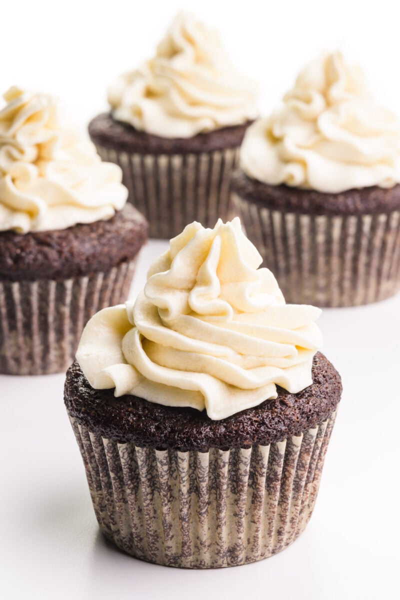 A cupcake has lots of swirly vegan ermine frosting on top. There are three more cupcakes with frosting in the background.