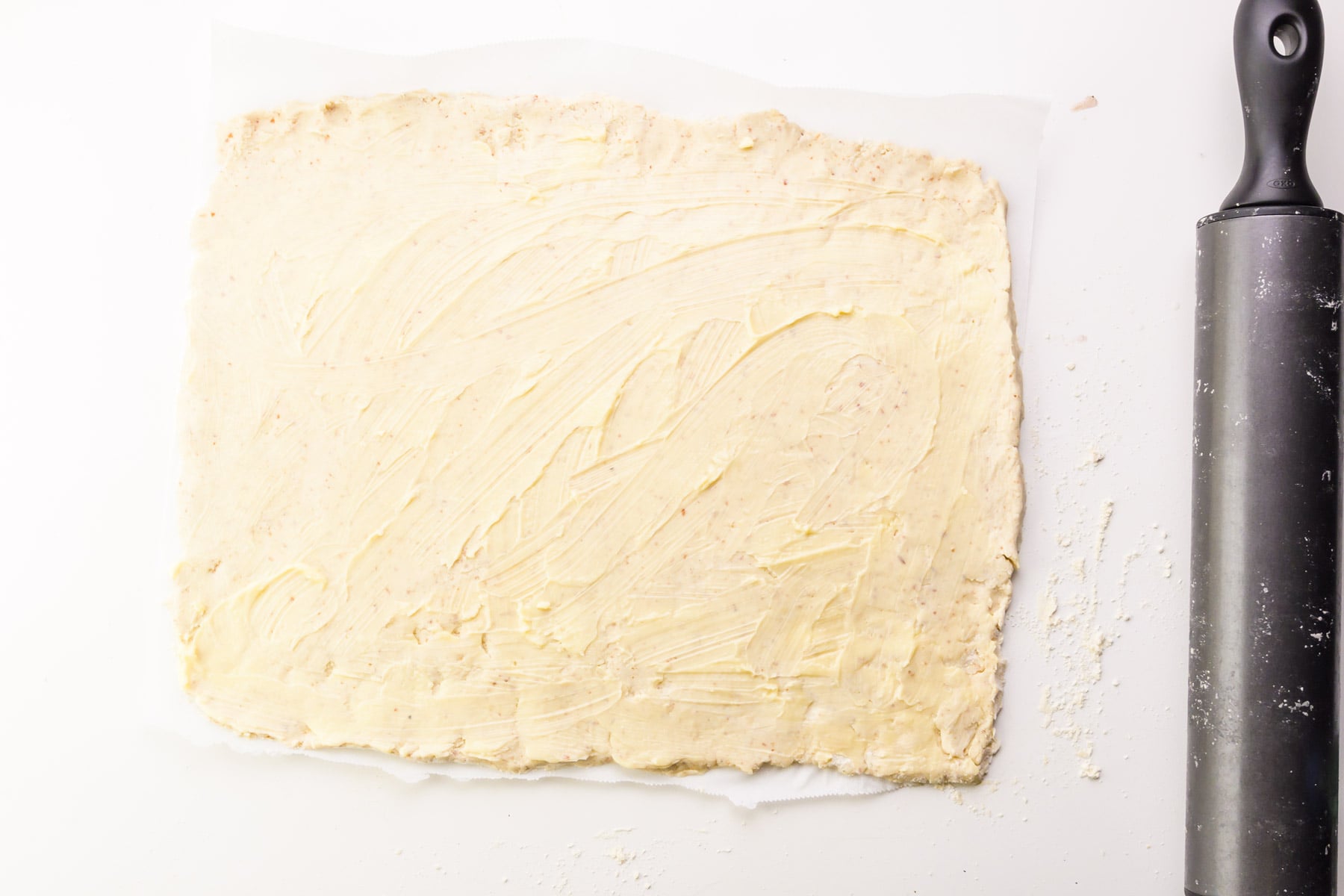 Rolled out dough has a layer of vegan butter spread on top. It sits beside a rolling pin.