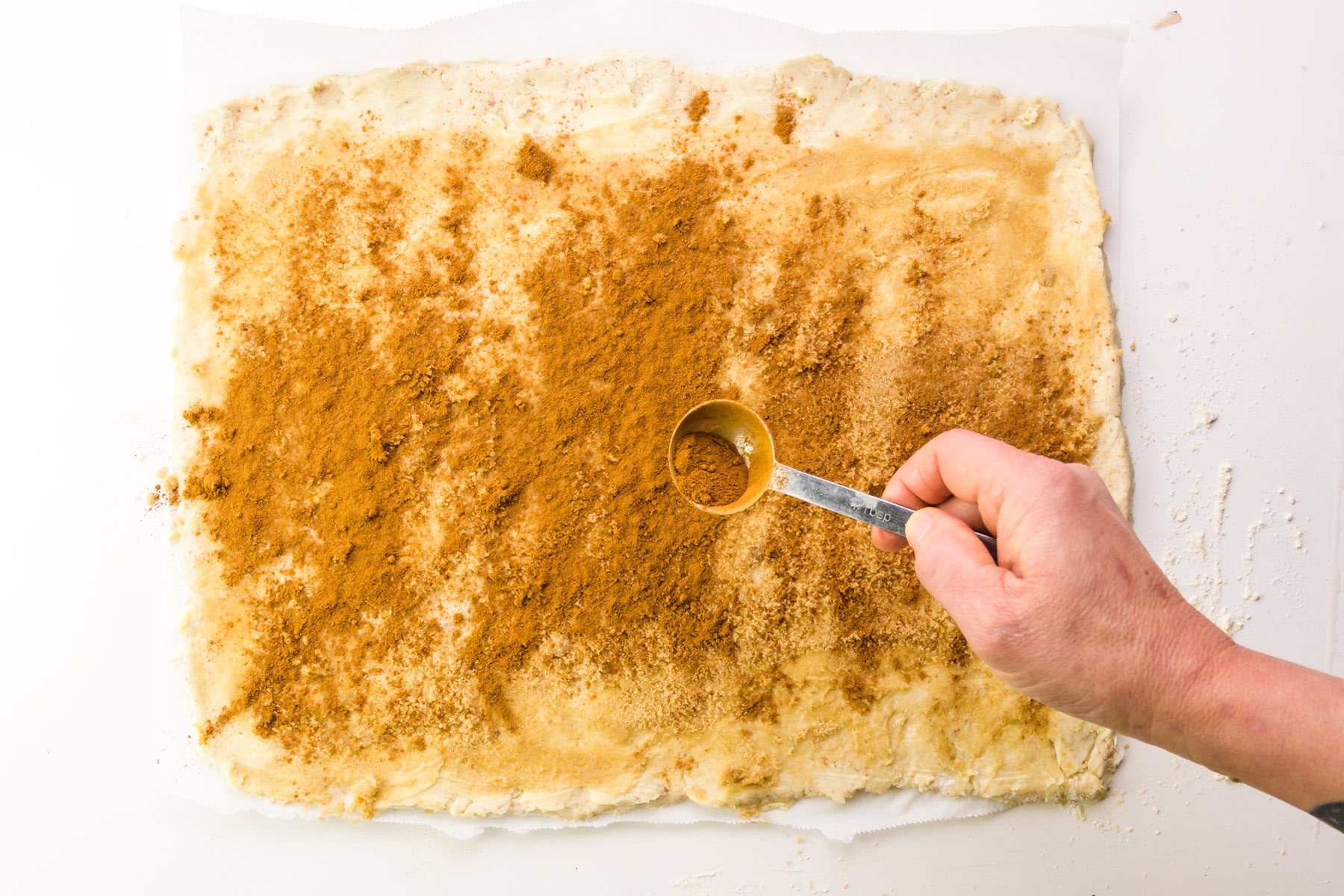 A hand holds a spoon sprinkling ground cinnamon over rolled out dough.
