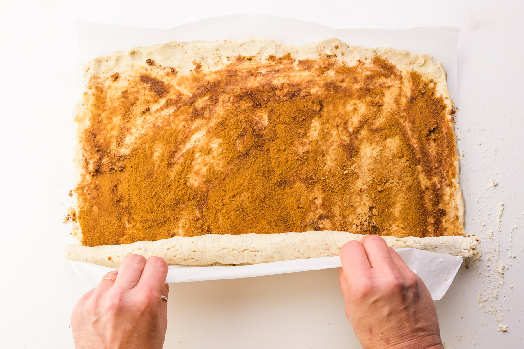 Two hands are using parchment paper to roll up dough into a log for cinnamon rolls.