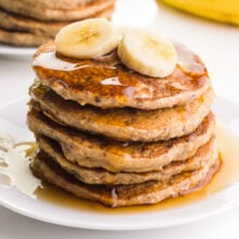 Sliced bananas are on top of a stack of pancakes. There are drizzles of syrup on the pancakes.