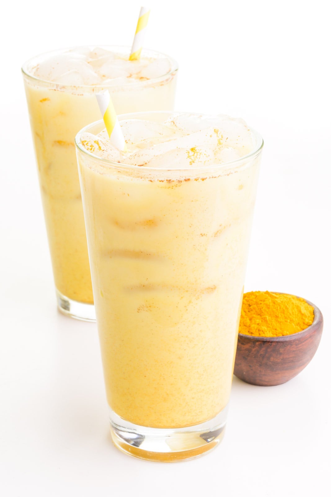 Two glasses of turmeric lattes sit next to a wooden bowl full of ground turmeric.
