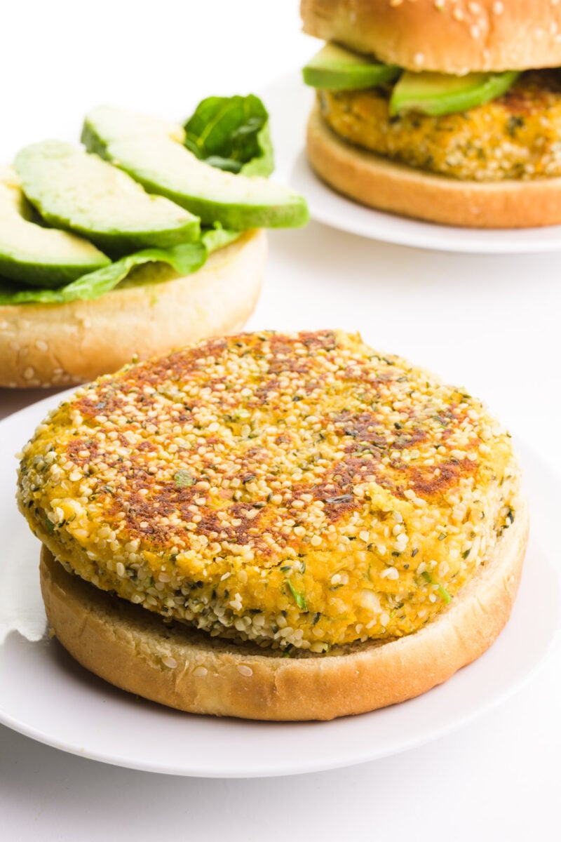 An open-face veggie burger is on a plate. The top side of the bun is behind it, with lettuce and avocado slices. There is a plate with another burger in the background.