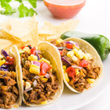 Three jackfruit tacos are on a plate with a jalapeño beside them. There is a bowl of salsa, a lime, and some fresh cilantro leaves in the background.