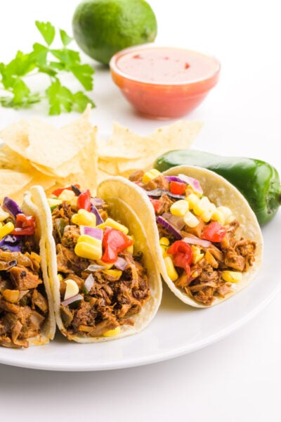 Three jackfruit tacos are on a plate with a jalapeño beside them. There is a bowl of salsa, a lime, and some fresh cilantro leaves in the background.