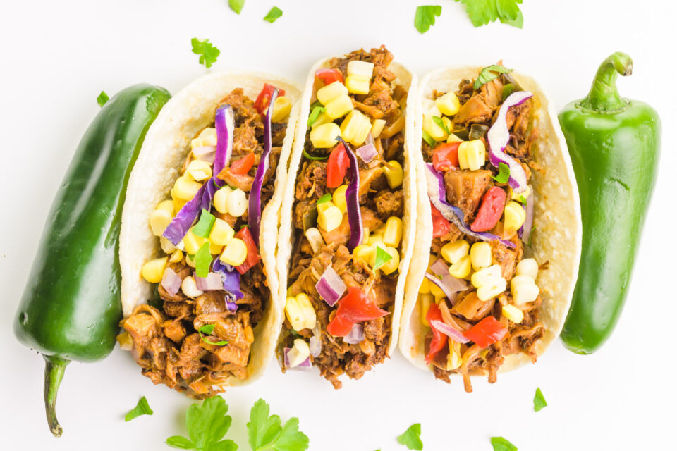 Looking down on 3 jackfruit carnitas tacos with jalapeños on both ends. Threre are bits of chopped cilantro around the tacos.