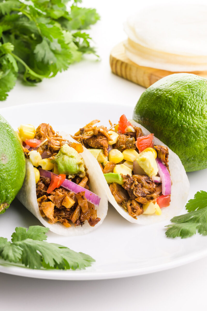 Two jicama tacos sit on a plate with limes and fresh cilantro. There is more cilantro and more jicama tacos in the background.