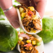 A hand holds a jicama taco. There's another taco in the background on a plate with limes and cilantro.