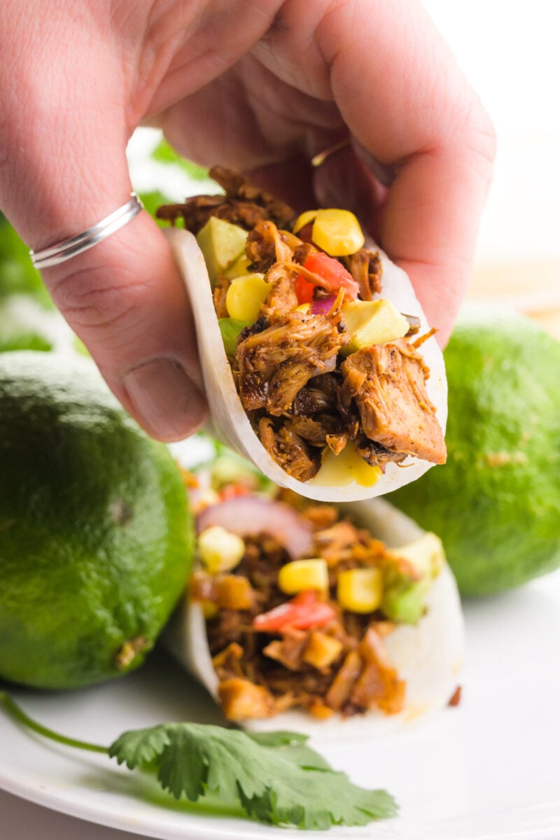 A hand holds a jicama taco. There's another taco in the background on a plate with limes and cilantro.