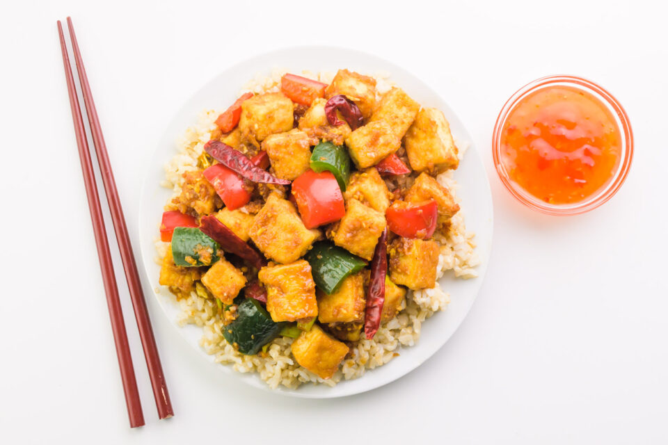 Looking down on a plate of tofu and vegetables. There is a bowl of sauce and chopsticks beside it.