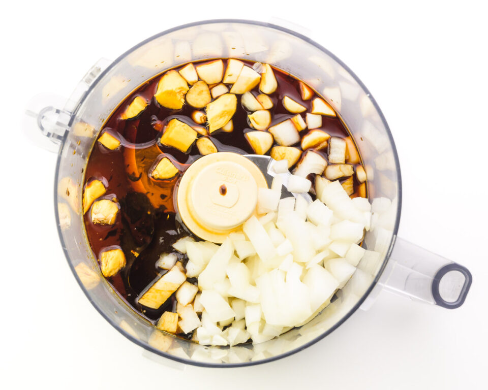 Ingredients like Tamari, chopped onions, garlic, and fresh ginger are in the bottom of a food processor bowl.