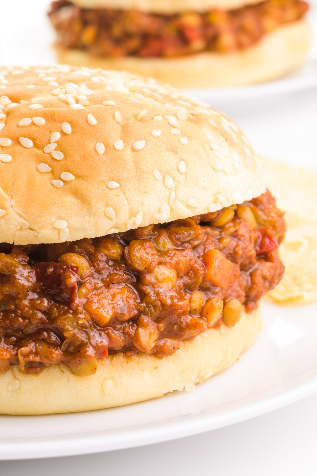 A bun holds lentil sloppy joes on a plate next to potato chips. There's another plate with the same sandwich in the background.