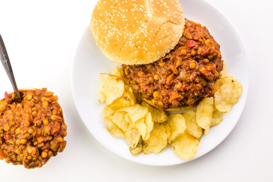 Looking down on a plate with a vegan sloppy Joe mixture on a bun, with the top bun to the side. There are potato chips on the plate, too. Next to the plate is a bowl with more of the vegan mixture with a spoon in in it.
