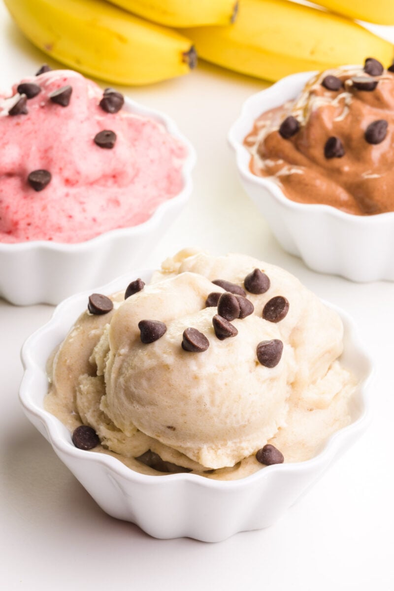 A bowl of nice cream sits in front of bowls of chocolate nice cream and strawberry nice cream. Bananas are in the background.