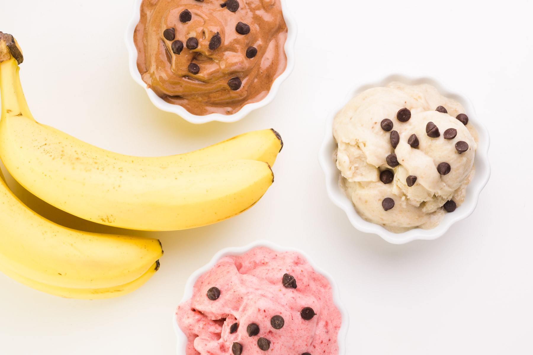 Looking down on three bowls of ice cream, one plain, one chocolate, and one strawberry. Bananas sit next to the bowls.