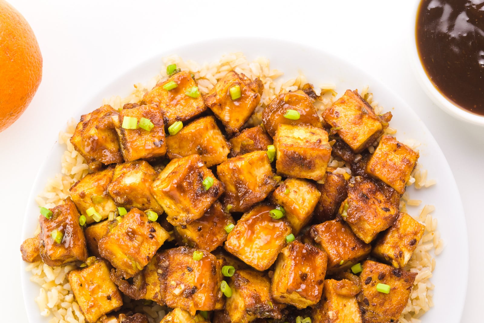 Looking down on a plate of tofu with sticky sauce on top. There's a bowl of sauce and an orange beside it.