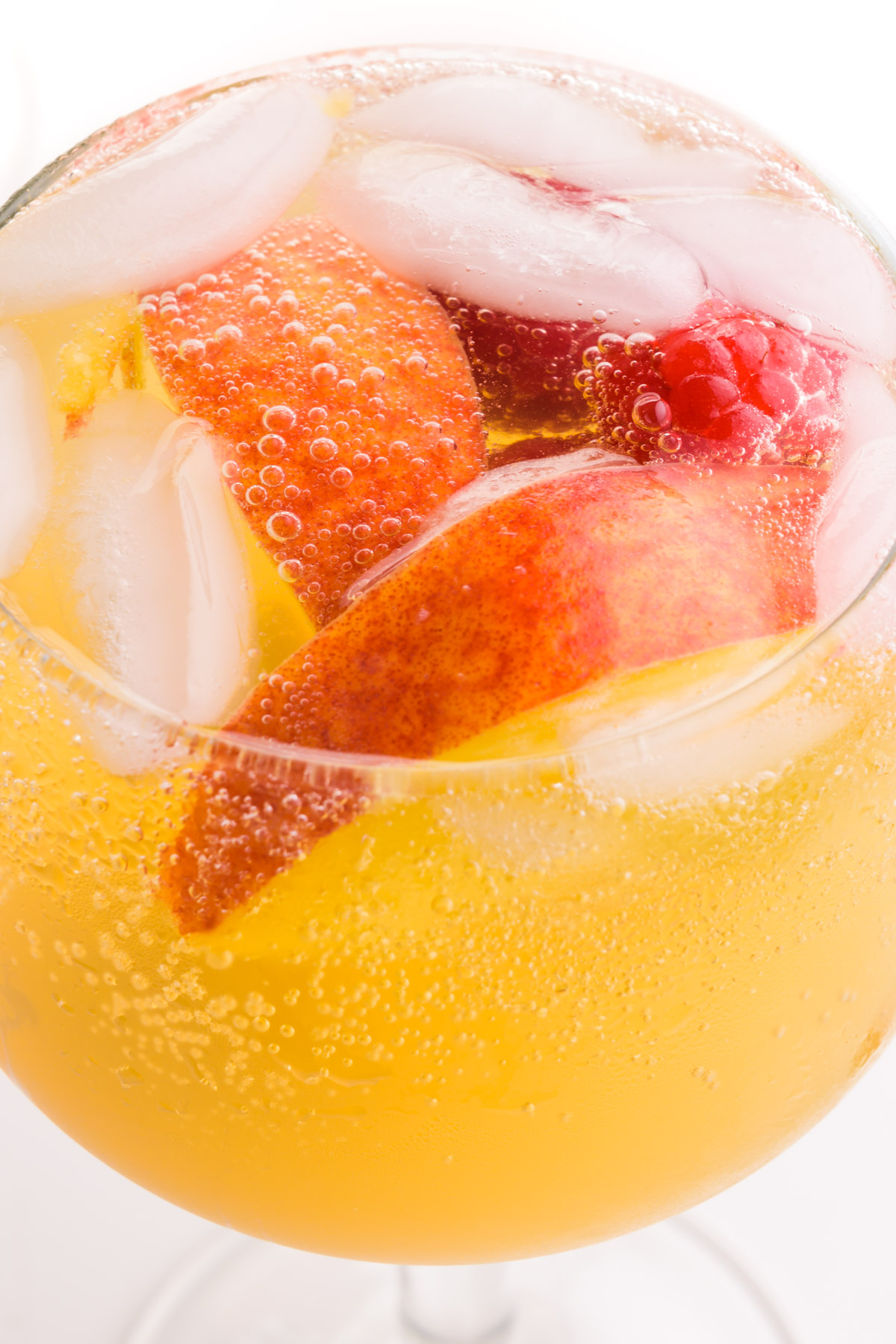 Looking into a wine glass full of peach sangria, ice cubes, raspberries, and peach slices.