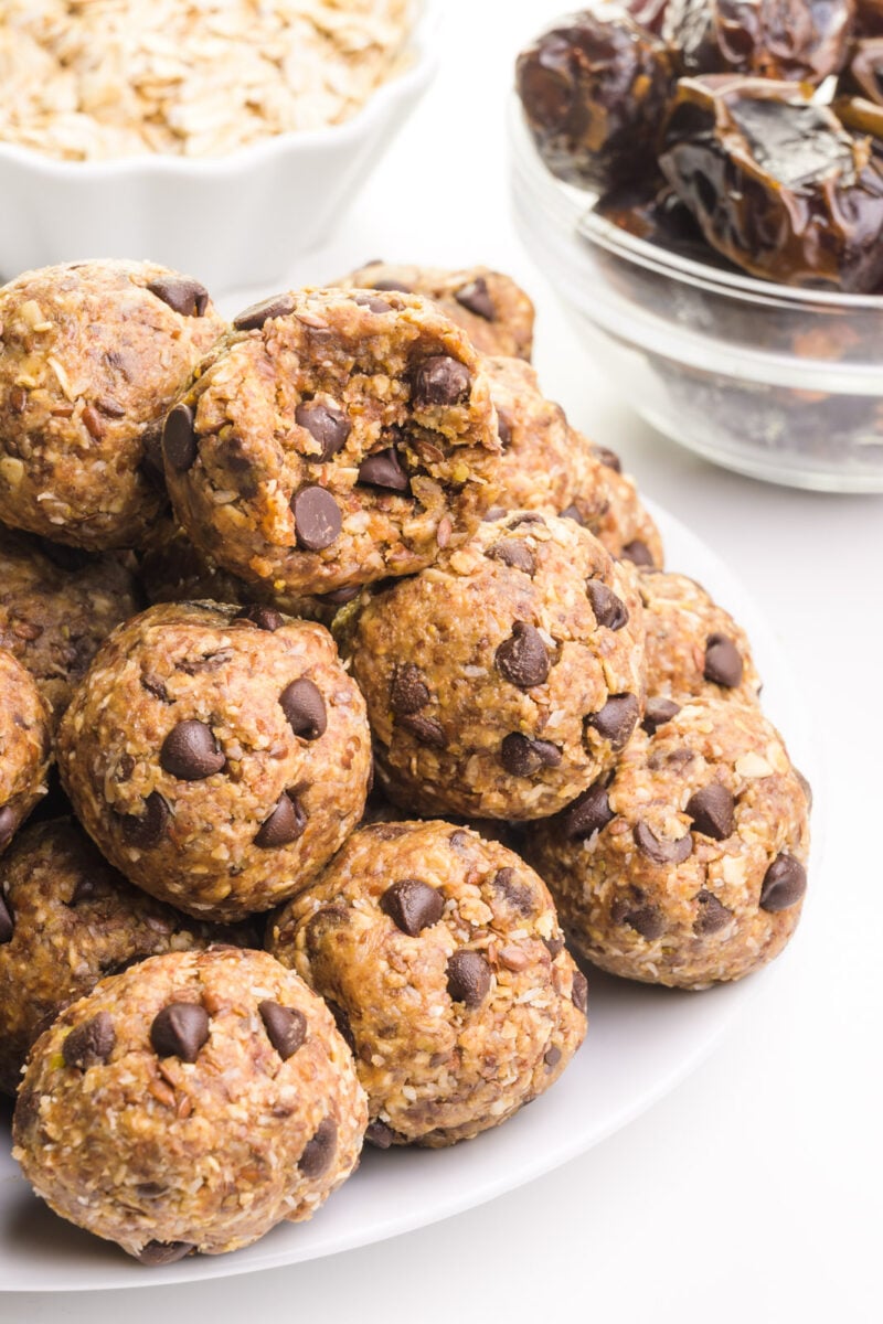 A mound of peanut butter energy balls sits on a white plate. The top ball has a bite taken out. There are two bowls in the background, one with dates and the other with oats.